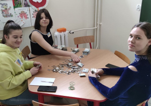Trzy dziewczynki siedzą przy stole i liczą pieniądze, które leżą na blacie.